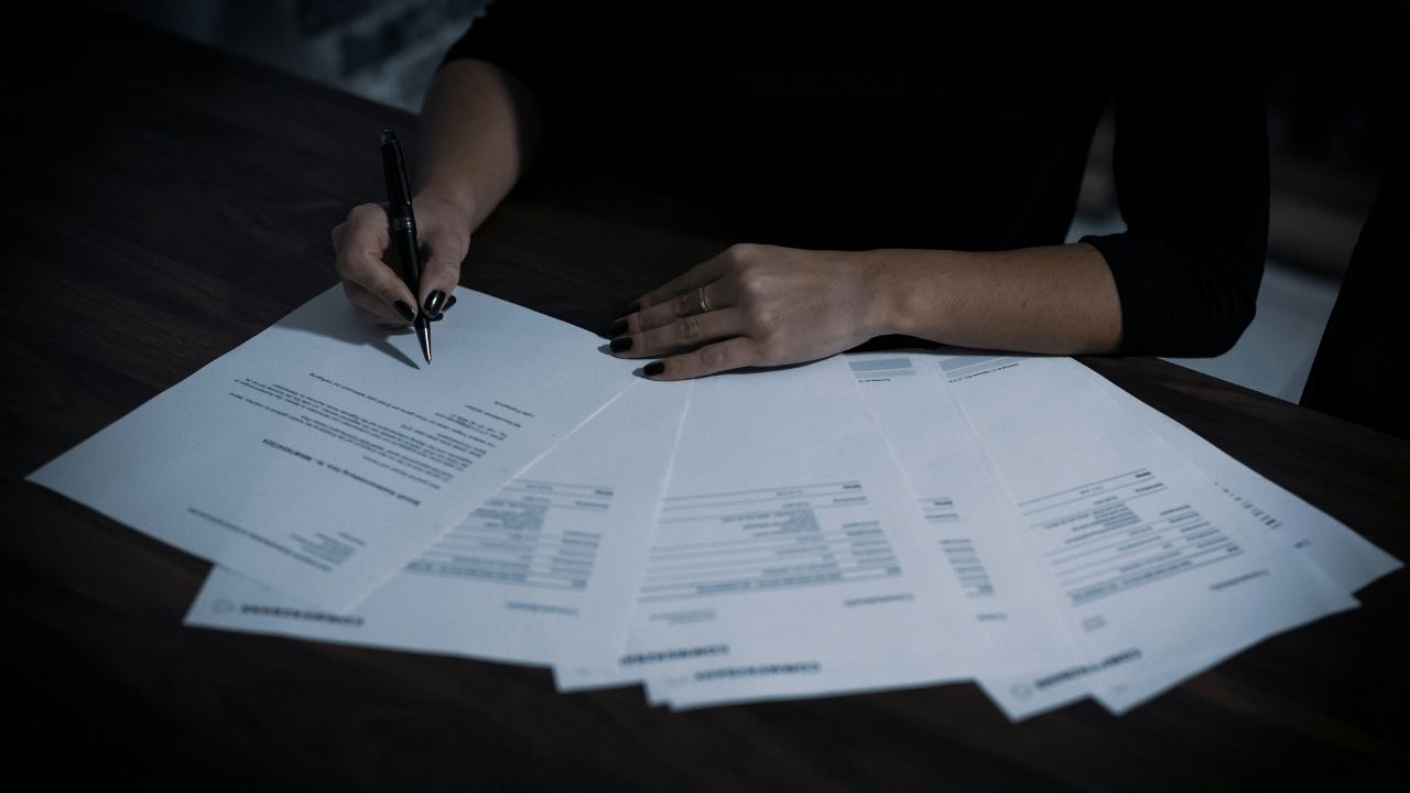 Several pages of a contract spread on the table whilst woman signs one page