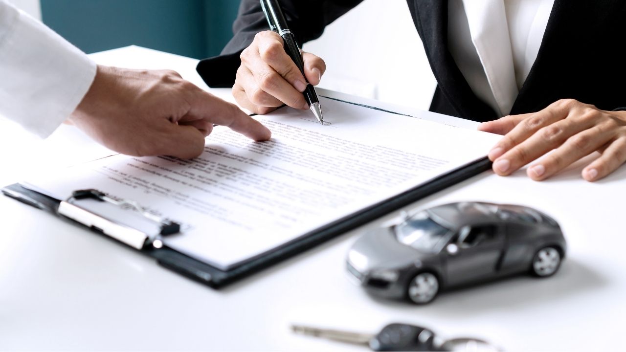 A man standing up and pointing to a specific line in a contract on a table and another man sitting on a chair whilst he signs the contract